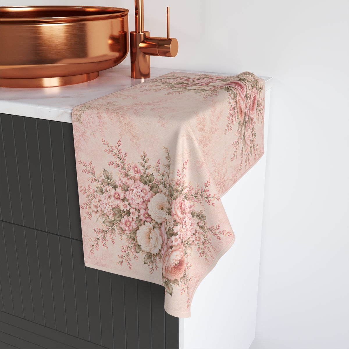 A rectangular hand towel with a vintage floral print in pink, beige, and green, draped over a white countertop next to a copper sink and faucet.