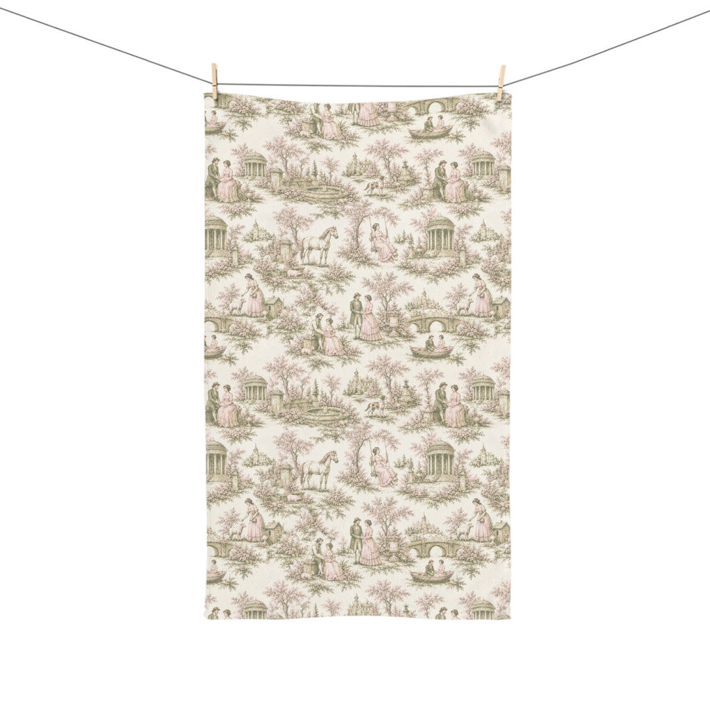 A rectangular hand or tea towel featuring a classic French toile de Jouy pattern with figures, trees, and classical architecture in muted pink and sage green on an off-white background, hanging from a clothesline with wooden clips.