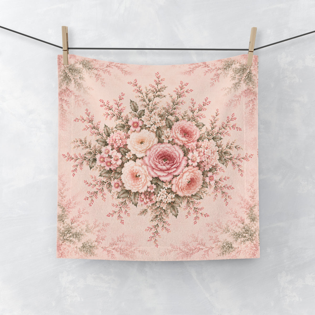A square washcloth hanging from two wooden clothespins on a black string against a light gray textured wall. The fabric features a vintage-style floral bouquet print in shades of pink, blush, cream, and muted green on a pale pink background.