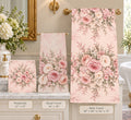 A coordinated set of floral bath towels displayed in a bathroom setting: a small washcloth, a hand towel, and a larger bath towel, all in a soft blush pink background with large rose bouquets and green foliage. Text labels show the sizes of each towel.
